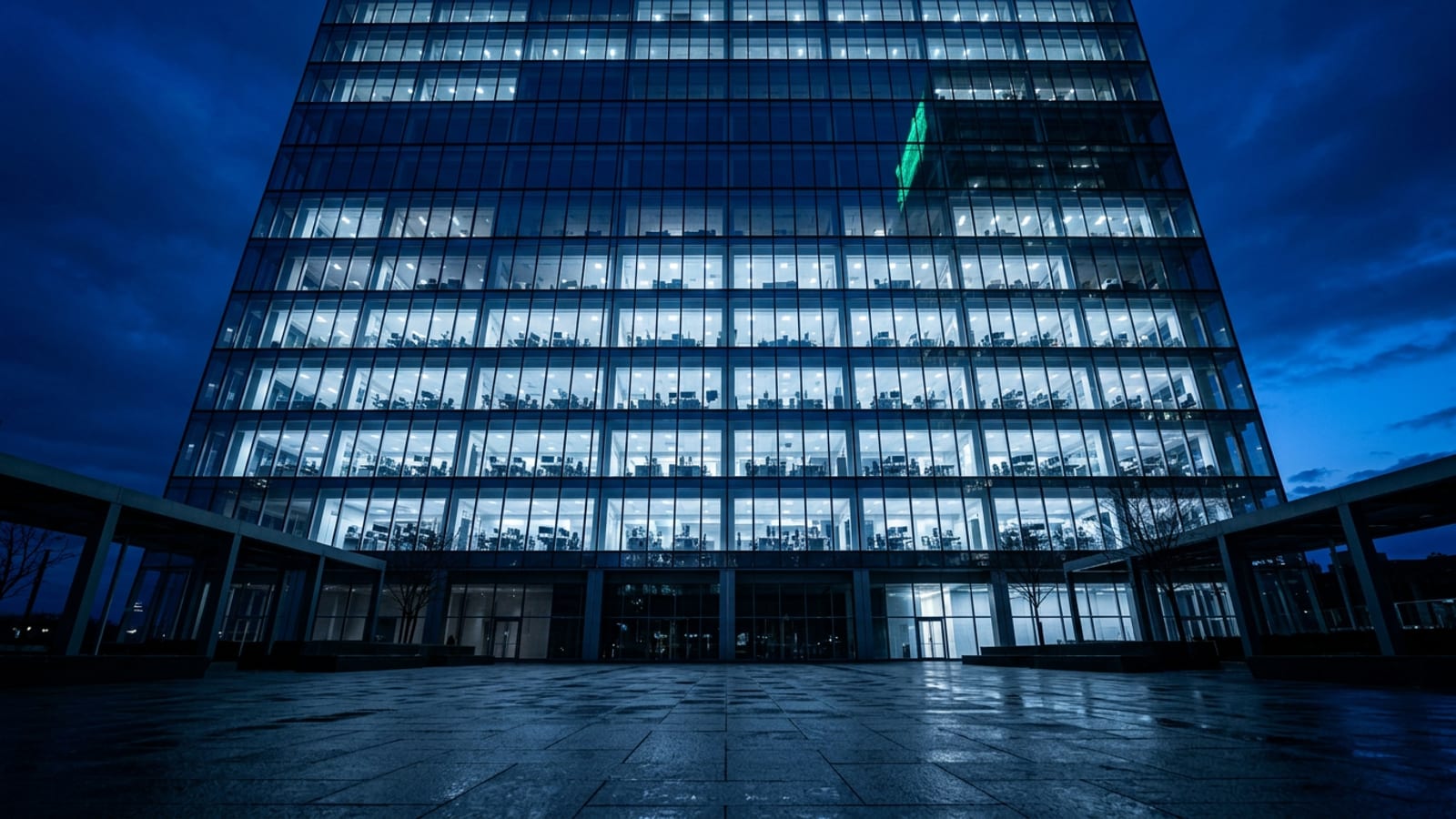 A Midtown Manhattan glass office tower at blue hour, its lit open-plan floors glowing through the facade