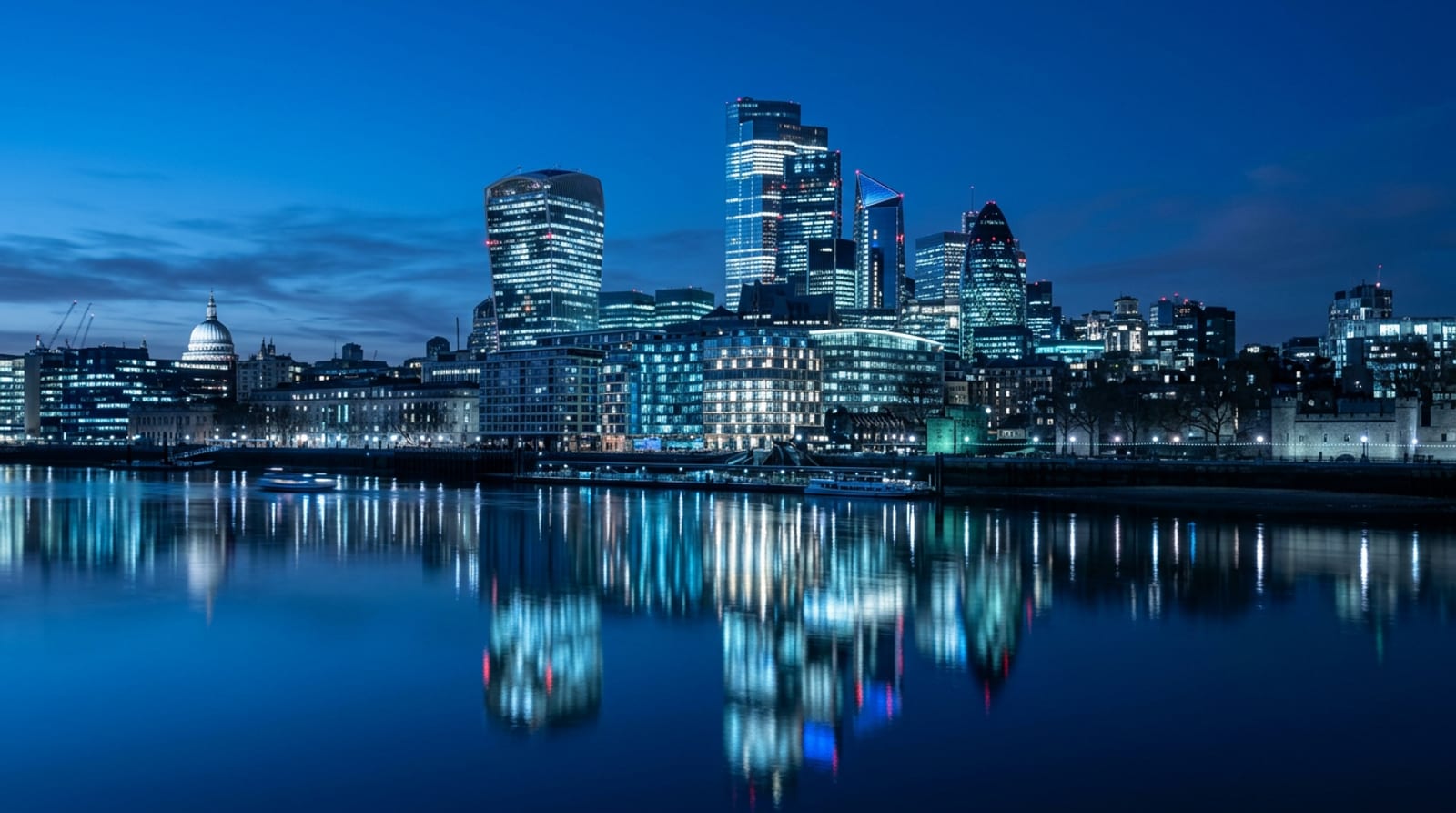 The City of London skyline at blue hour seen across the Thames, glass towers with lit windows reflected in the river