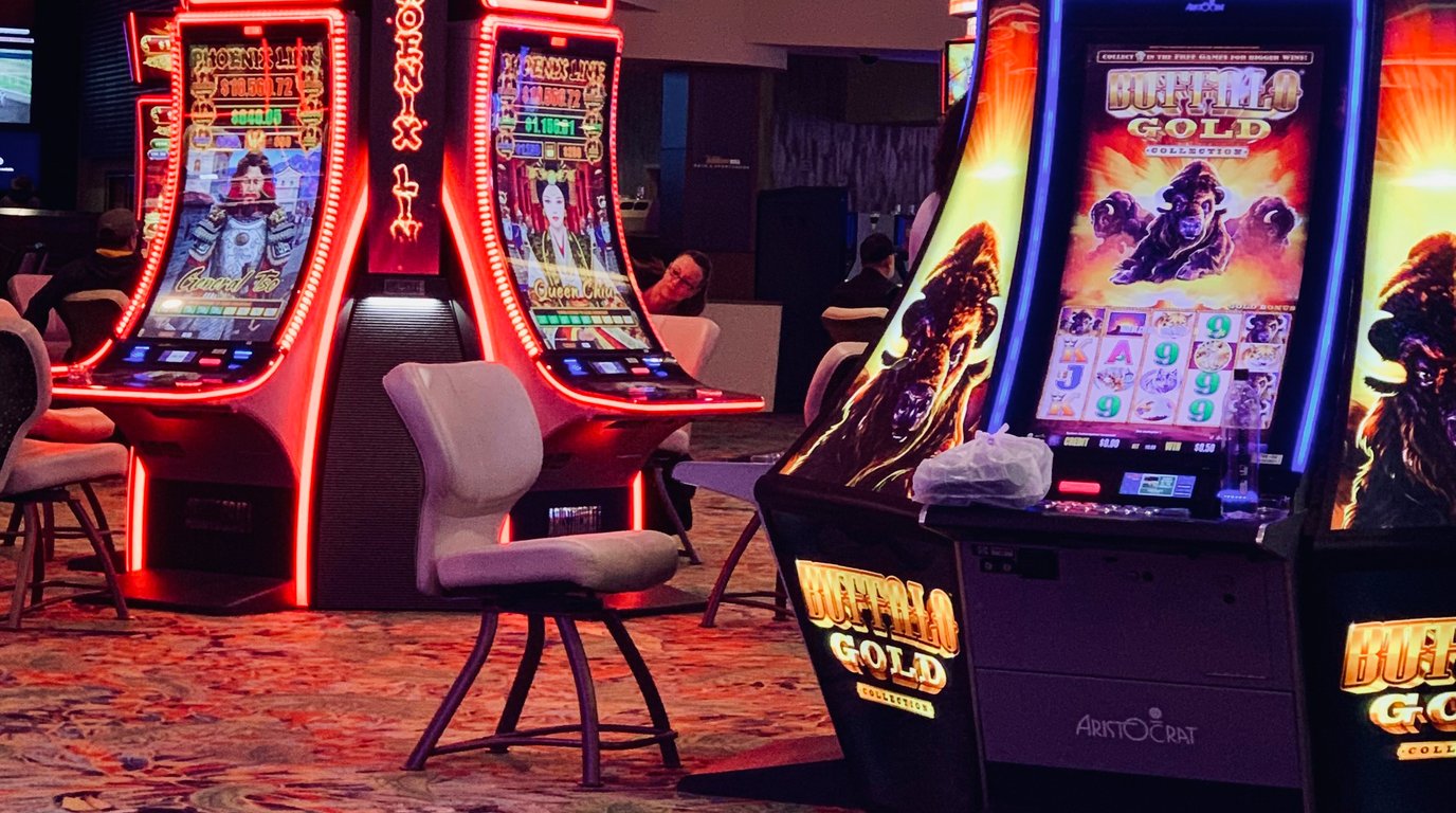 A wide view of a gaming compliance testing floor where technicians inspect both physical slot machines and banks of monitors running online casino game diagnostics.