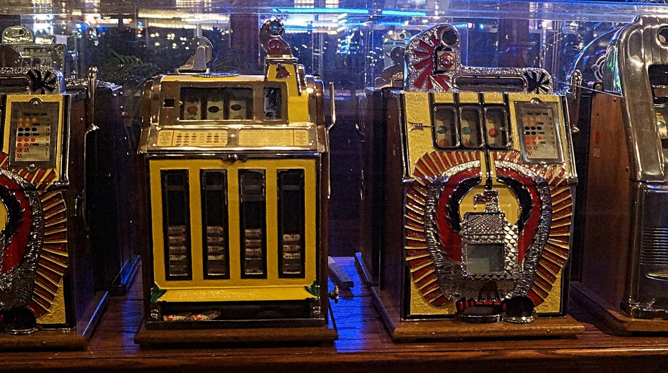 A gaming compliance auditor with a tablet walking past both a row of physical slot machines and a bank of monitors displaying online casino game testing dashboards.
